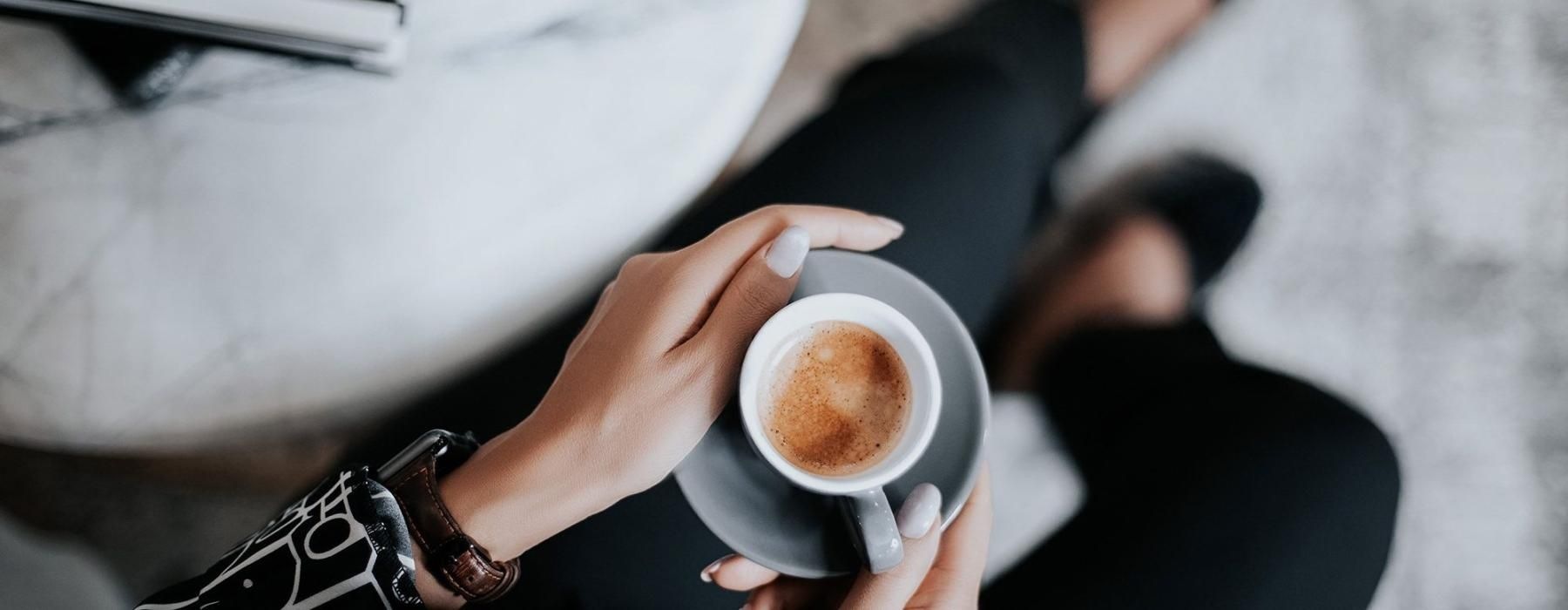 business woman sits next to a marble table with books and holds a saucer with a cup of espresso