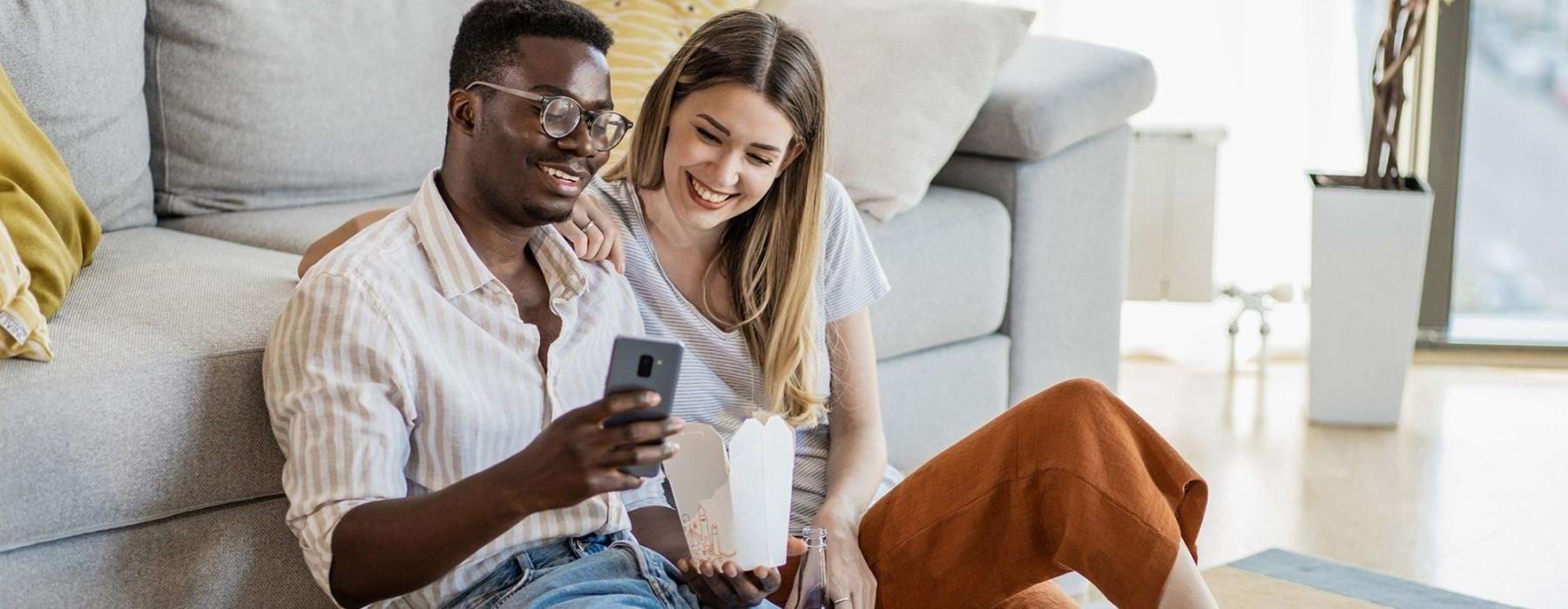 a man and woman with take out, sit against a couch on their living room floor and watch at their cell phone